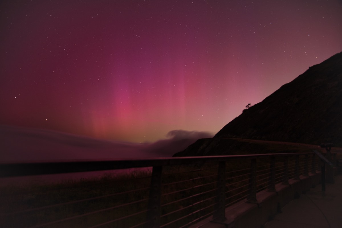 Pink and magenta aurora over a coastal overlook, with a fog bank rolling in below a dark headland