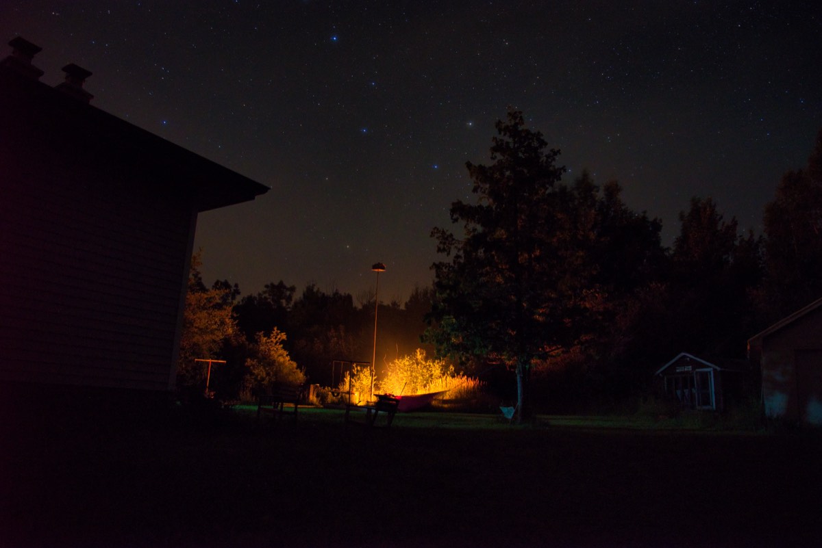 A lamplit yard at night with a hammock strung between trees under a star-filled sky